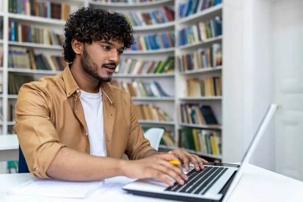 Estudiante latino sonriente consulta información en su portátil en la biblioteca de la universidad