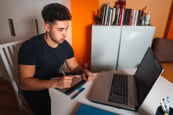 Joven estudiante toma apuntes junto a su portátil mientras planifica su formación en ciberseguridad desde casa